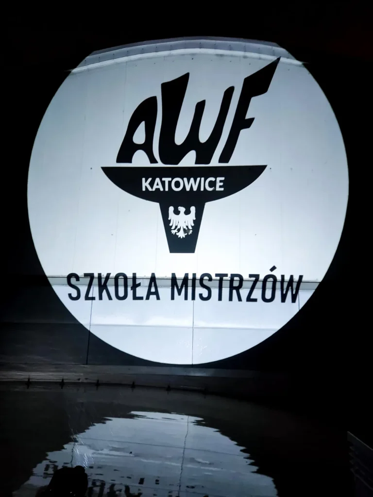 Logo AWF Katowice — Szkoła Mistrzów wyświetlone projektorem GOBO na ścianie